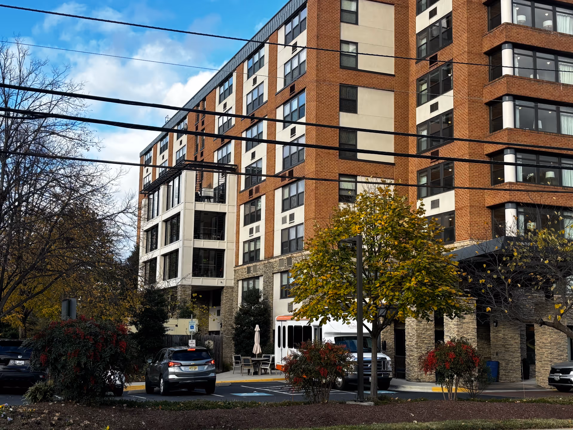 A multi-story brick building with a covered entrance, trees, parked cars and a shuttle bus in front.