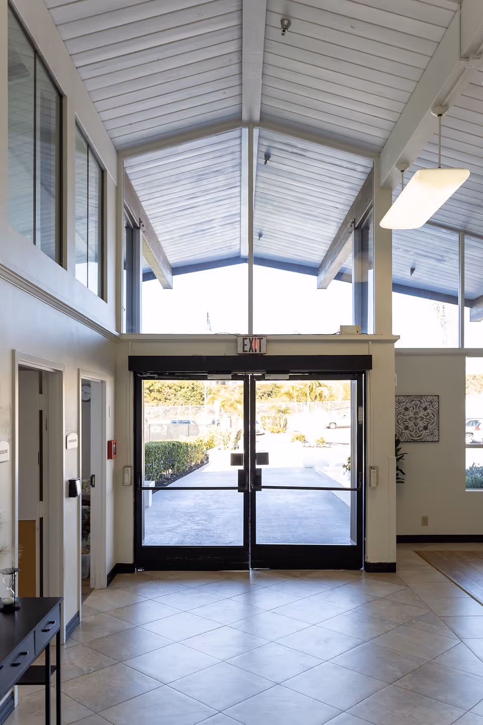 Interior view of an entrance area with double glass doors leading outside. The ceiling is vaulted with large windows above the doors allowing natural light to fill the space. There is a tiled floor and a small table with decorative items on the left side.