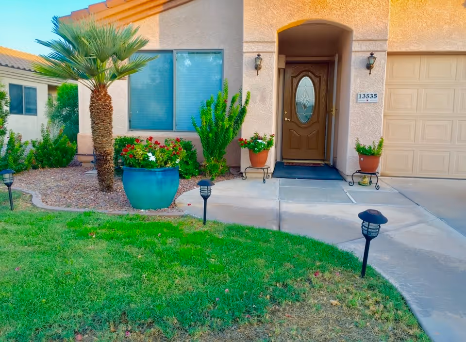 Front entrance of a stucco single-story house with a brown decorative door, potted plants, a palm tree, and a green lawn.