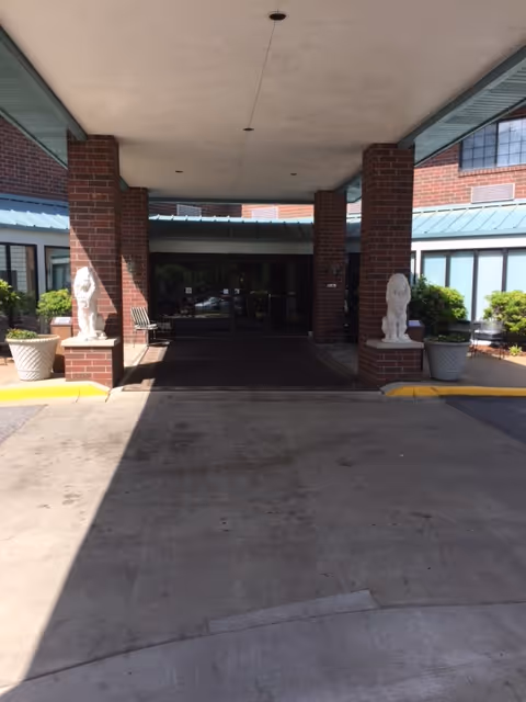 Covered entrance area of a building with brick pillars and two white lion statues on pedestals, potted plants on either side, and glass doors leading inside.