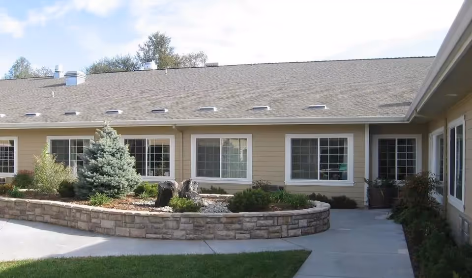 Single-story memory care building exterior with a raised stone planter, shrubs and multiple large windows facing a paved courtyard.