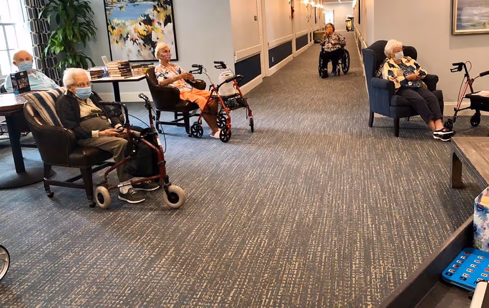 A hallway in an assisted living facility with elderly residents sitting in chairs and using walkers and wheelchairs. The hallway has carpeted flooring, light-colored walls, and doors along the sides. There is a table with books and a large painting on the wall.