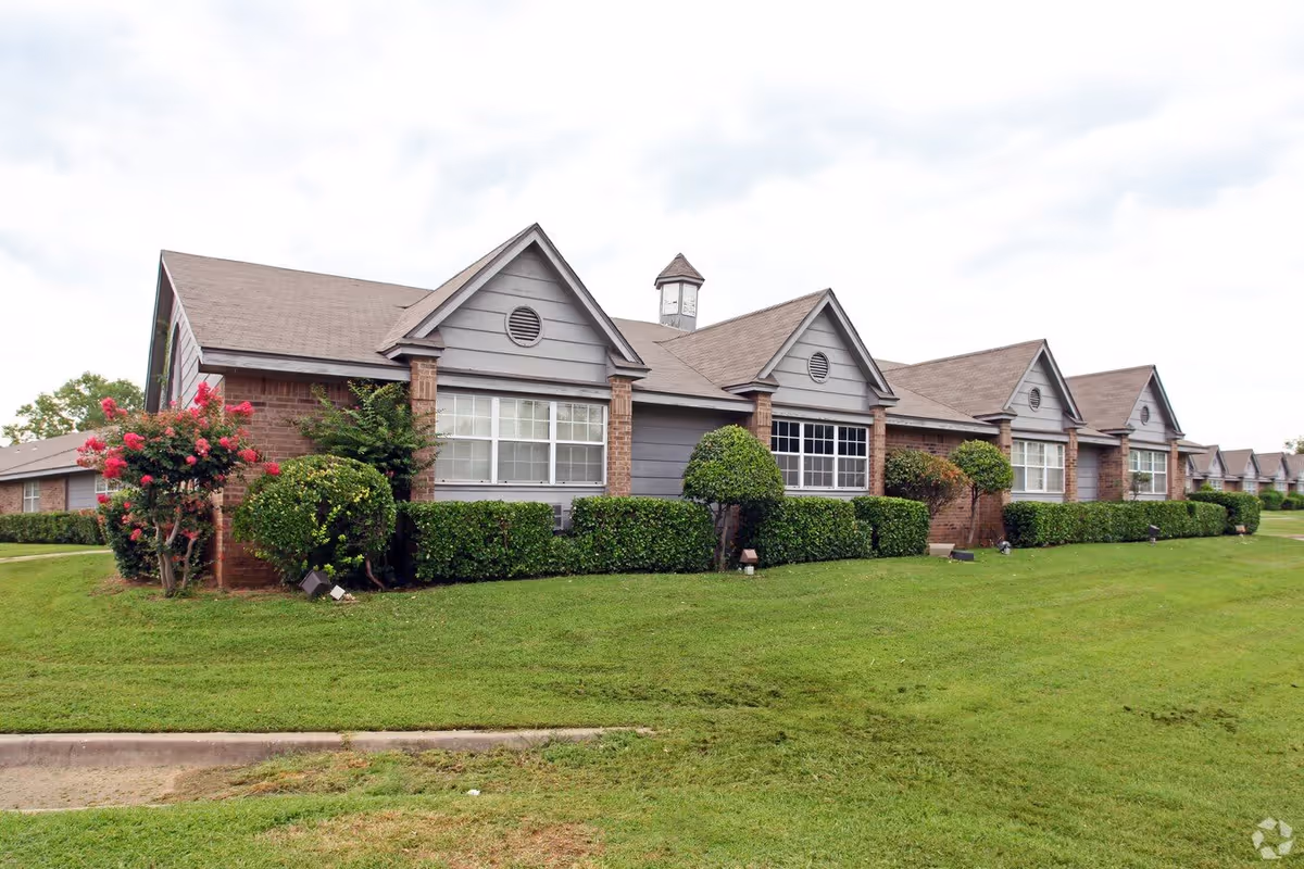 Exterior view of a single-story assisted living facility building with brick and gray siding, multiple windows, neatly trimmed bushes, and a well-maintained green lawn under a cloudy sky.