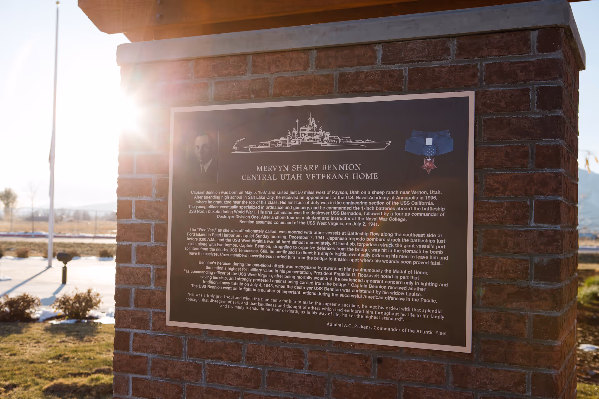 A commemorative plaque mounted on a brick pillar outdoors at Central Utah Veterans Home. The plaque honors Mervyn Sharp Bennion, detailing his military service and heroism during World War II, accompanied by an illustration of a battleship, a portrait of Bennion, and a Medal of Honor image. The sun is shining brightly from the left side, casting a lens flare.