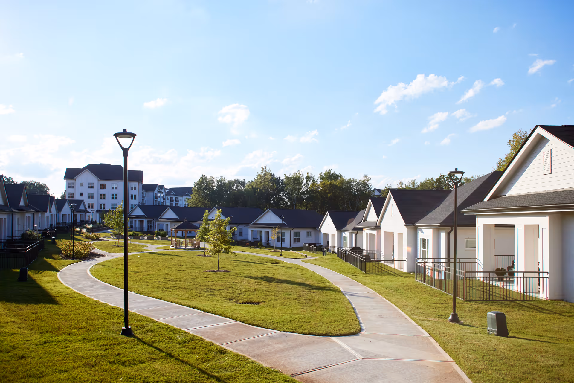 Sunlit courtyard with a winding sidewalk, lamp posts, and rows of single-story cottages with a larger building in the background.