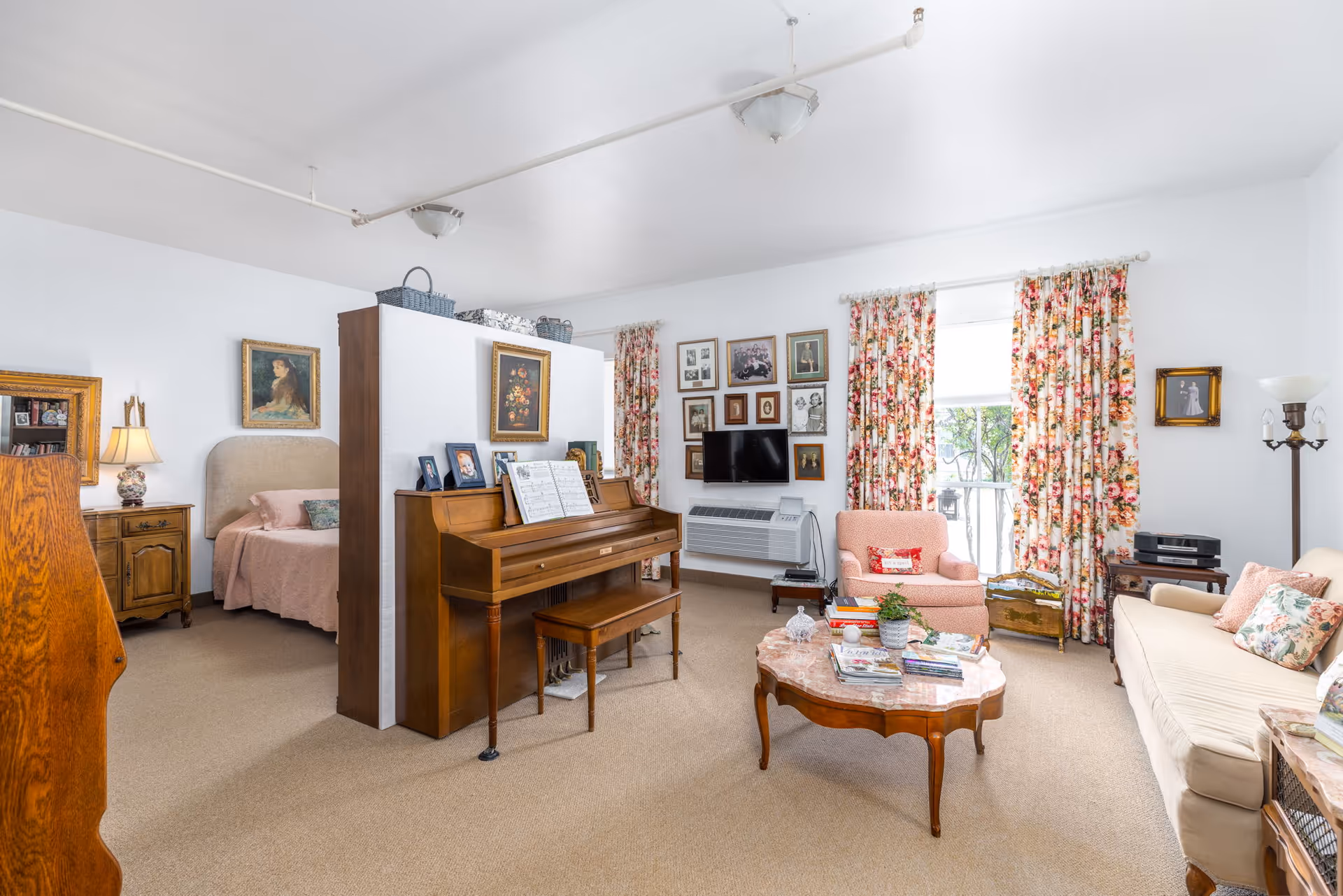 A bright and cozy living area with floral curtains, a beige sofa with floral pillows, a pink armchair, a wooden coffee table with magazines and plants, and a wooden piano against a partial wall. Behind the partial wall is a bedroom area with a bed, nightstand, lamp, and framed artwork on the walls. The living area also features a wall-mounted TV surrounded by framed photos and a window letting in natural light.