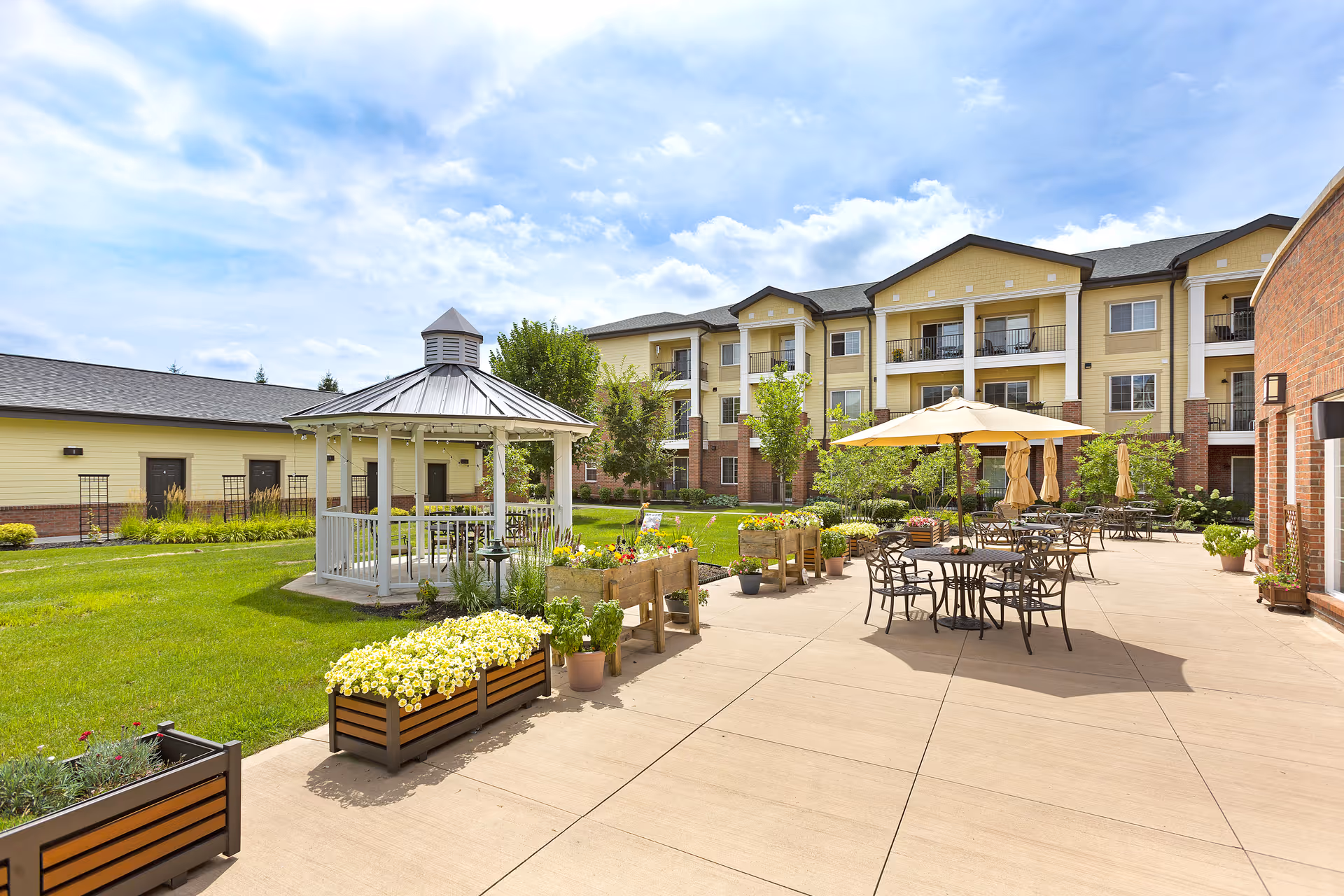 Outdoor patio area at StoryPoint Powell featuring metal tables and chairs with umbrellas, surrounded by flower planters and a white gazebo on a grassy lawn. The background shows a three-story residential building with balconies and a partly cloudy sky.