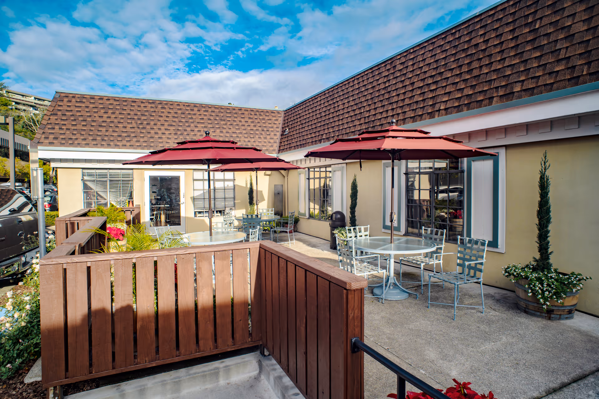 Outdoor patio area at South Marin Health & Wellness Center with round glass tables, metal chairs, and red umbrellas. The patio is enclosed by a wooden fence and surrounded by potted plants and shrubs. The building has beige walls and brown shingled roof under a partly cloudy blue sky.