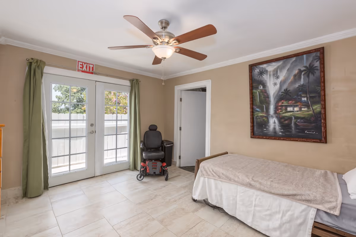 A bedroom in an assisted living facility with a single bed covered with a beige blanket, a ceiling fan with wooden blades, a large framed painting of a waterfall and tropical scenery on the wall, a black mobility scooter near a door, and double glass doors with green curtains leading outside.