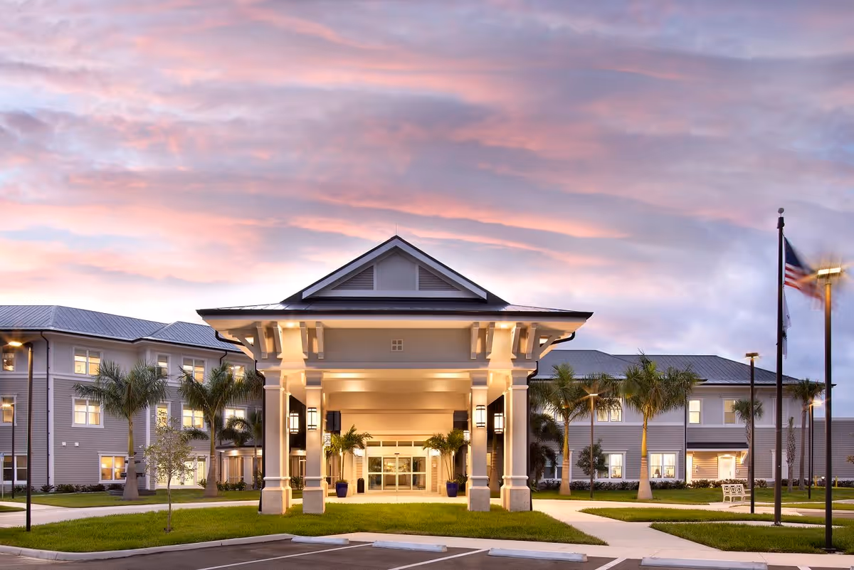 Front entrance of a senior living building with a covered porte-cochère, palm trees, and an American flag under a colorful sunset sky.
