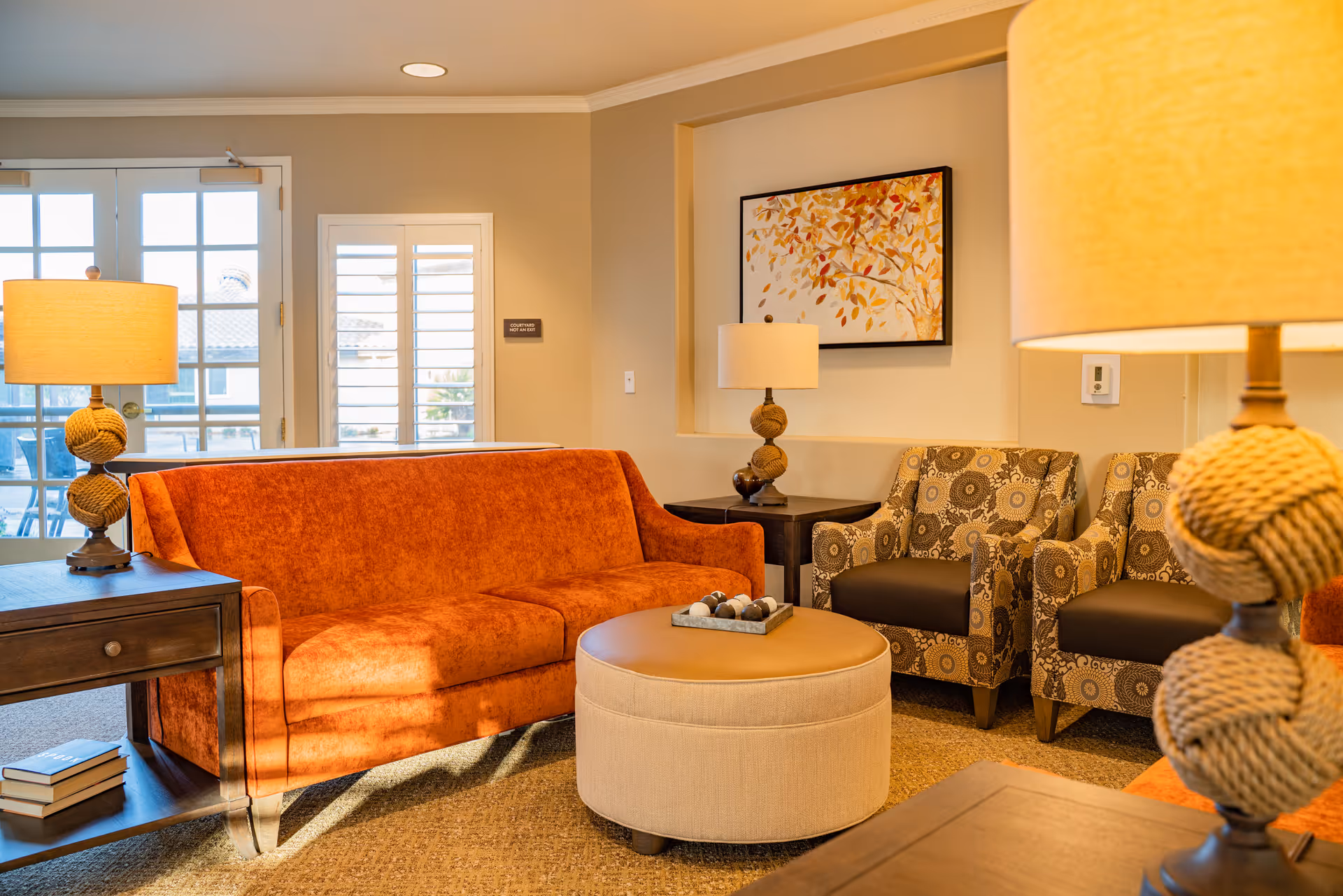 A cozy living room area with an orange sofa, two patterned armchairs, a round beige ottoman, two side tables with lamps, and a framed artwork of autumn leaves on the wall. French doors and a window with white shutters are visible in the background.