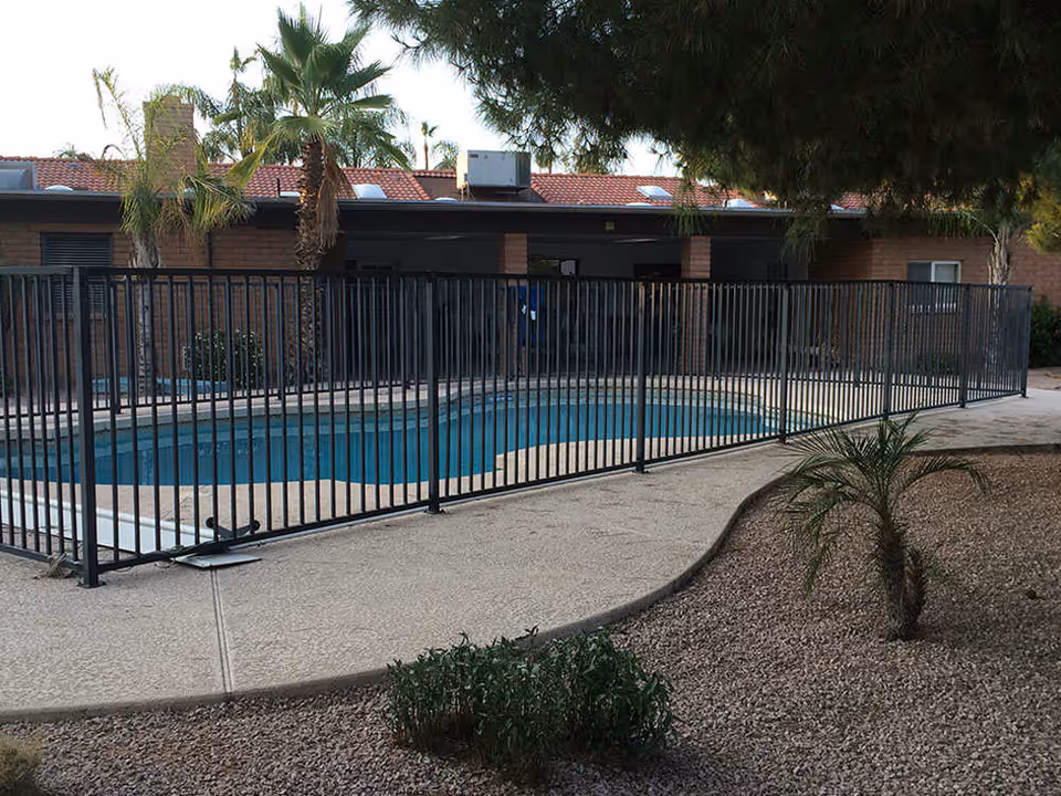 Outdoor swimming pool enclosed by a black metal fence with a concrete walkway surrounding it. There are palm trees and other plants around the pool area, and a single-story building with a red tile roof in the background.
