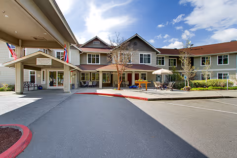 Front entrance of a two-story senior living building with a covered drive-up, outdoor seating, and American flags.