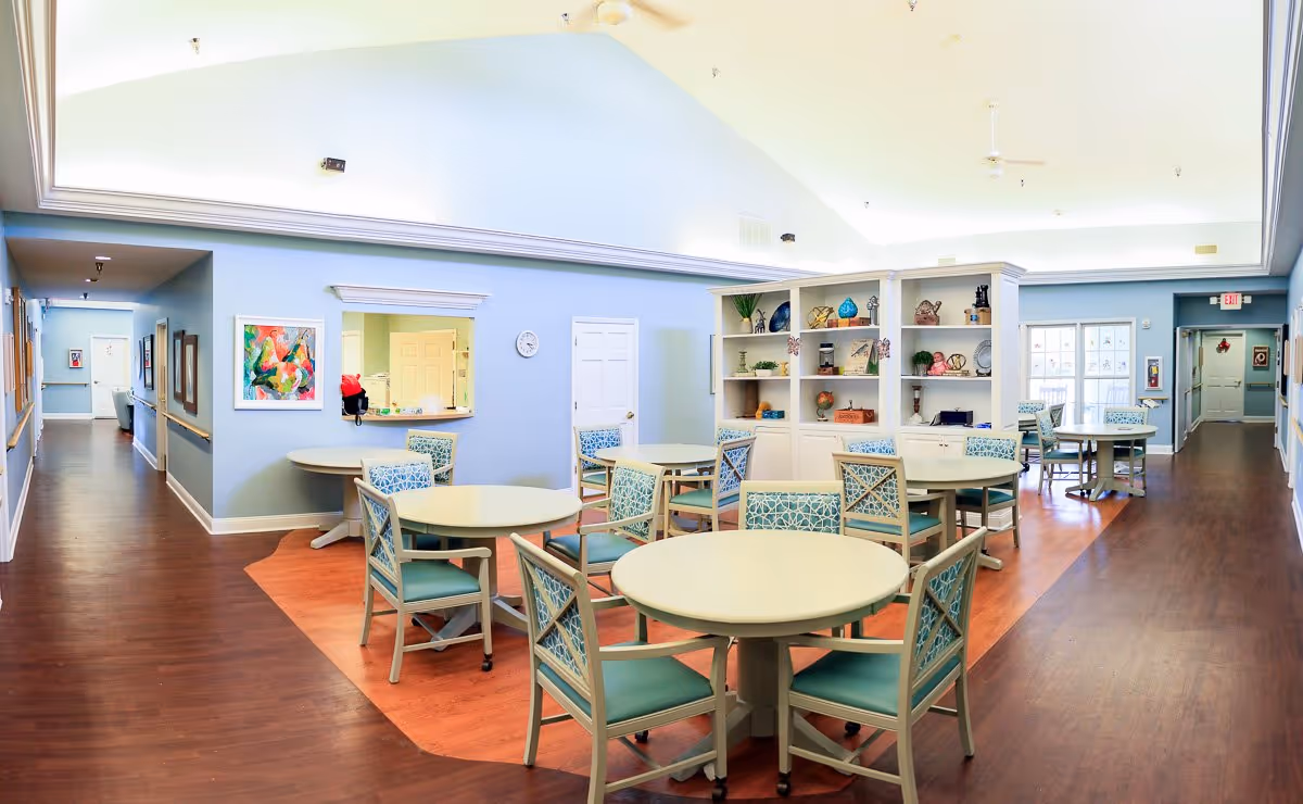 Bright communal dining and activity room with round tables, patterned chairs, shelving and a vaulted ceiling.