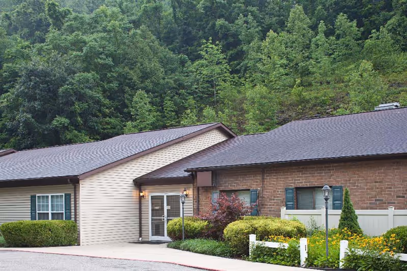 Exterior view of a single-story senior living facility building with beige siding and brick walls, surrounded by green bushes, flowers, and trees in the background.