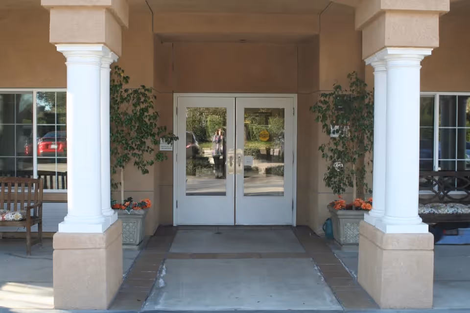 Entrance to a building with double glass doors framed by two white columns. There are potted plants with orange flowers on either side of the doors, wooden benches with cushions on both sides, and windows reflecting the outside surroundings.