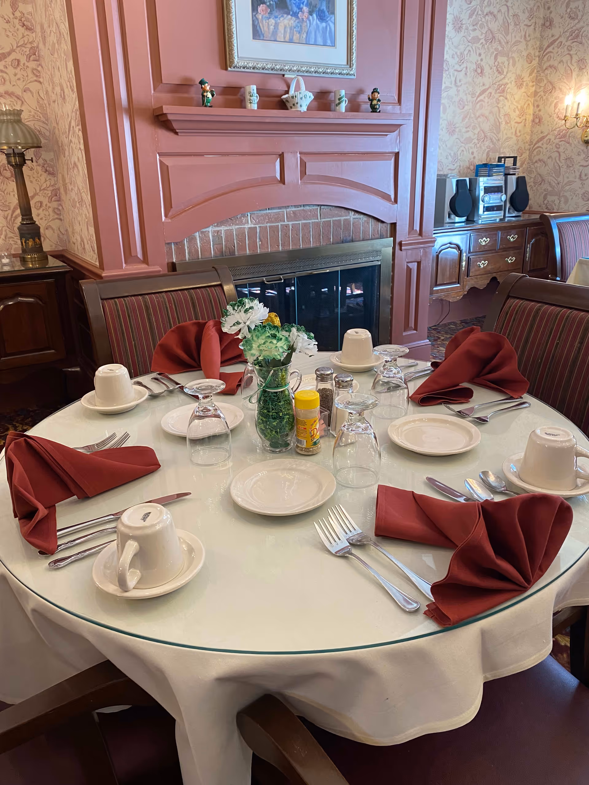 A round dining table set for four with white plates, upside-down cups on saucers, silverware, and folded maroon napkins. A small vase with green and white flowers and condiments are in the center. The table is in a room with a pink fireplace, floral wallpaper, and wooden furniture including a sideboard with a stereo system.