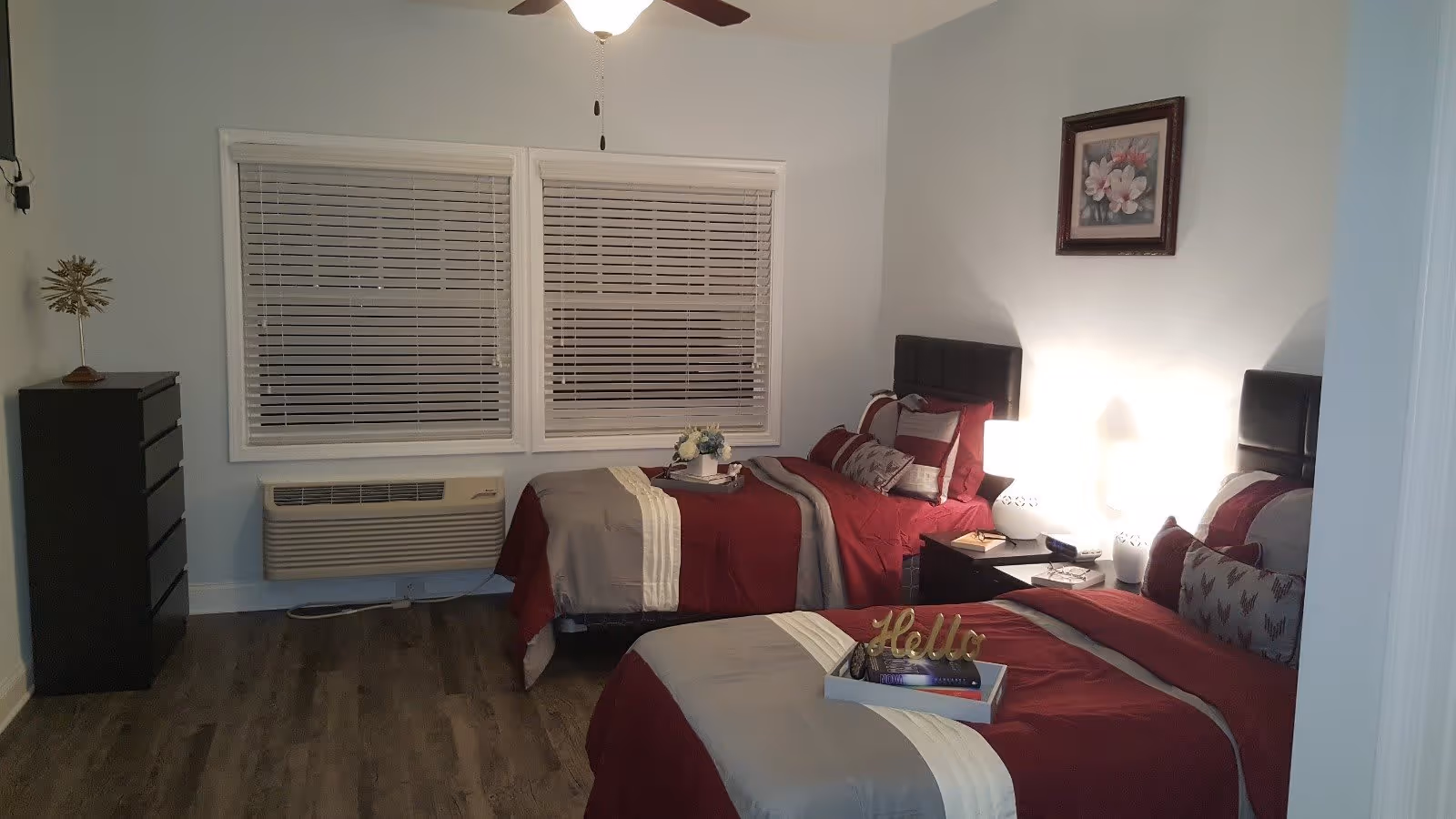 A bedroom with two single beds dressed in red and beige bedding. Each bed has multiple pillows and a tray with decorative items on it. There is a nightstand with two white lamps between the beds, a framed floral picture on the wall, a black dresser on the left side, and a window with closed blinds above a wall-mounted heater.