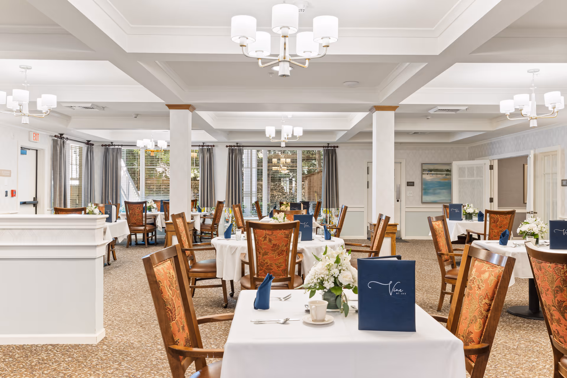A bright and elegant dining room with multiple round tables covered in white tablecloths. Each table is set with blue napkins, menus, and floral centerpieces. The chairs have wooden frames with orange patterned upholstery. Large windows with gray curtains allow natural light to fill the room. The ceiling features white coffered beams and modern chandeliers.