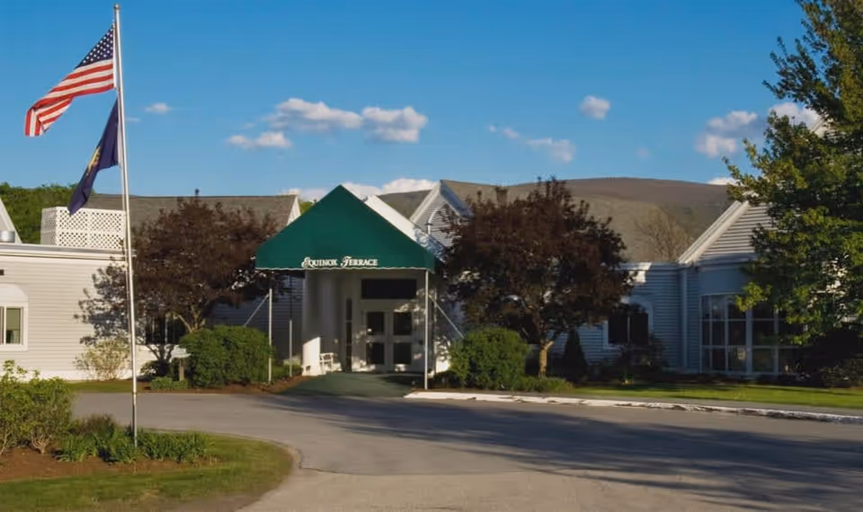 Exterior view of Equinox Terrace facility with a green entrance canopy displaying the facility name. The building is white with multiple windows, surrounded by trees and bushes. Two flagpoles with the American flag and another flag are visible on the left side under a blue sky with scattered clouds.