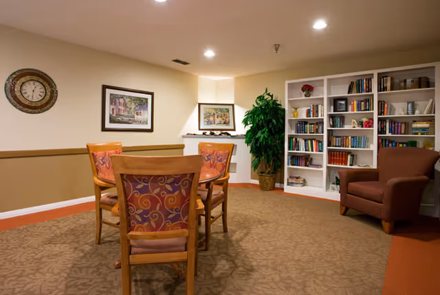 A cozy interior room with a round wooden table surrounded by four chairs with patterned cushions. There is a brown armchair to the right, a large white bookshelf filled with books and decorative items, a tall green plant in a basket, and framed artwork on the walls. A clock is mounted on the left wall.