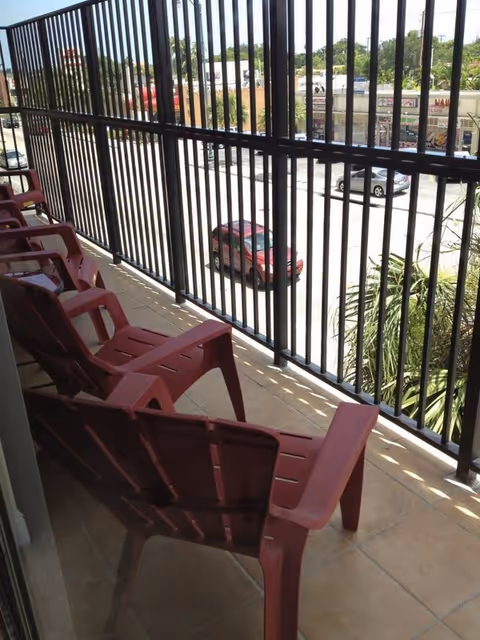 A balcony with several red plastic chairs lined up facing outward. The balcony has a black metal railing and overlooks a street with parked cars and some greenery.