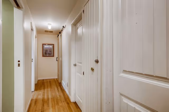 A narrow indoor hallway with wooden flooring and white paneled doors on both sides. The walls are painted white, and there is a framed picture hanging at the end of the hallway under a ceiling light.