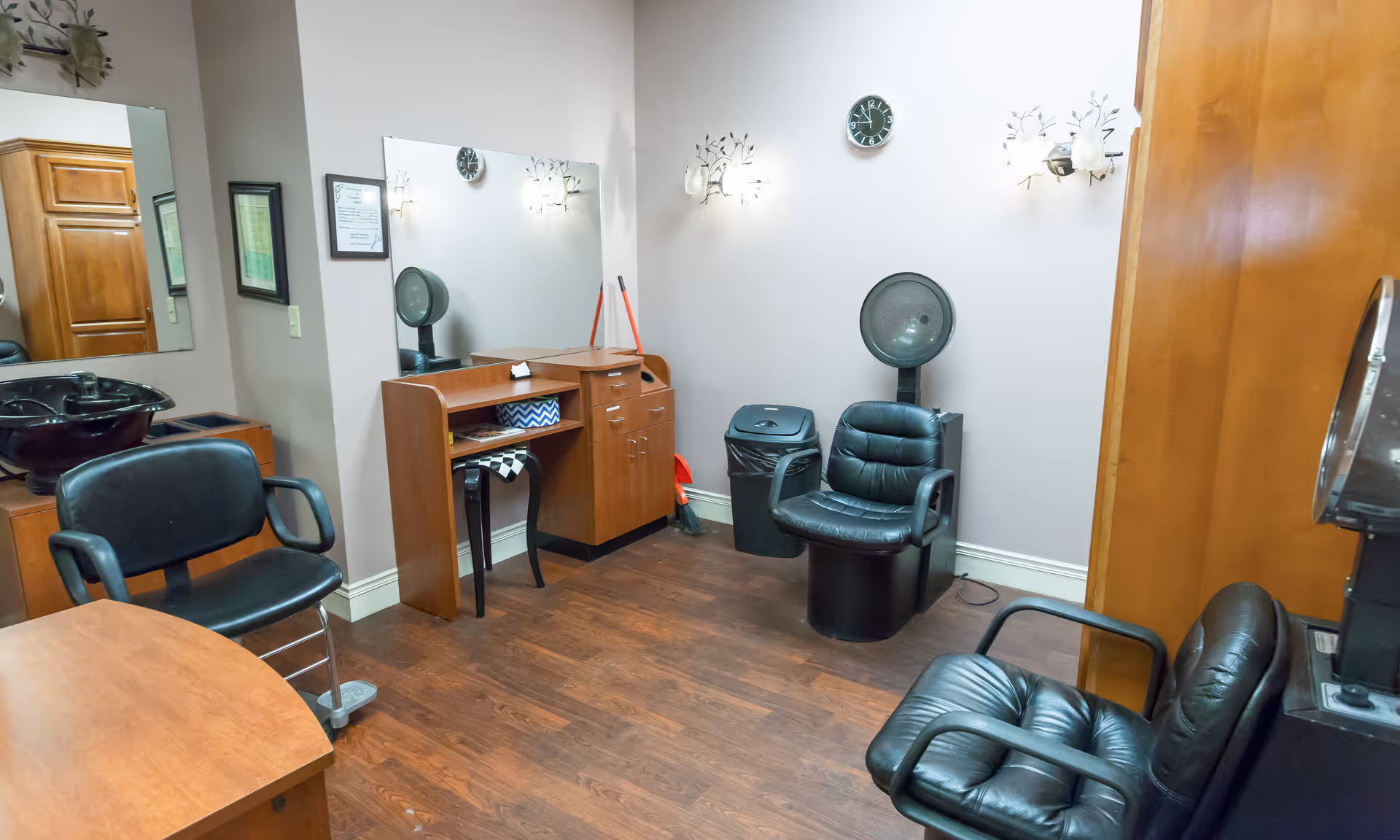 Interior salon room with styling chairs, a hooded hair dryer, mirrors, and wooden cabinets on wood flooring.
