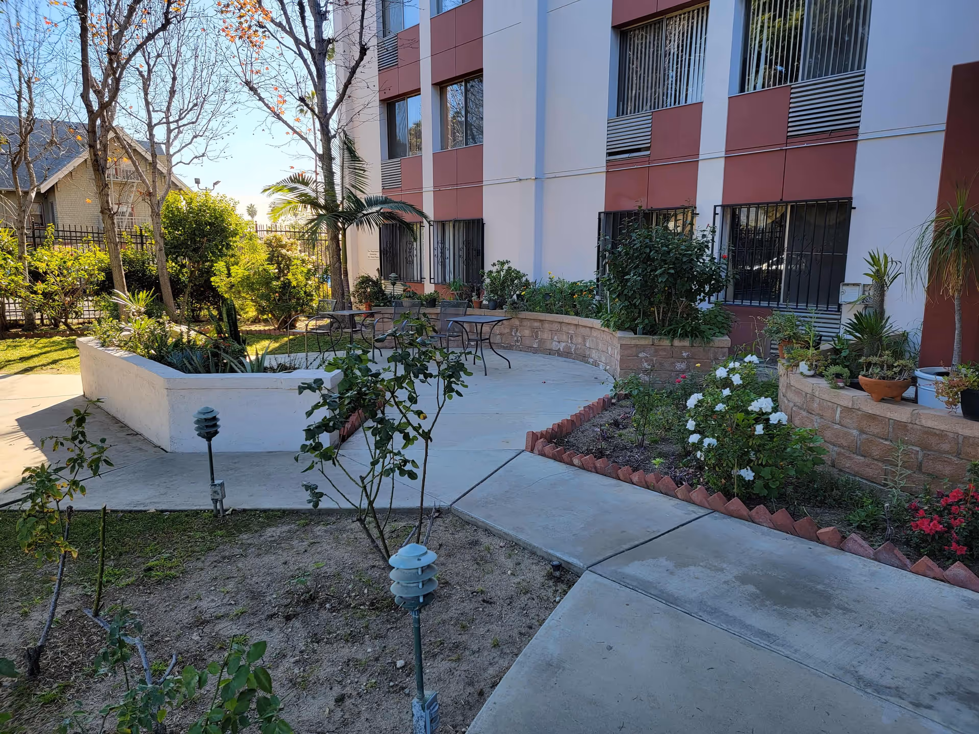 Outdoor garden area with paved walkways, various plants, and flowers. There are a few metal tables and chairs near the building with a red and white exterior. Trees and shrubs surround the area, and the sky is clear and blue.