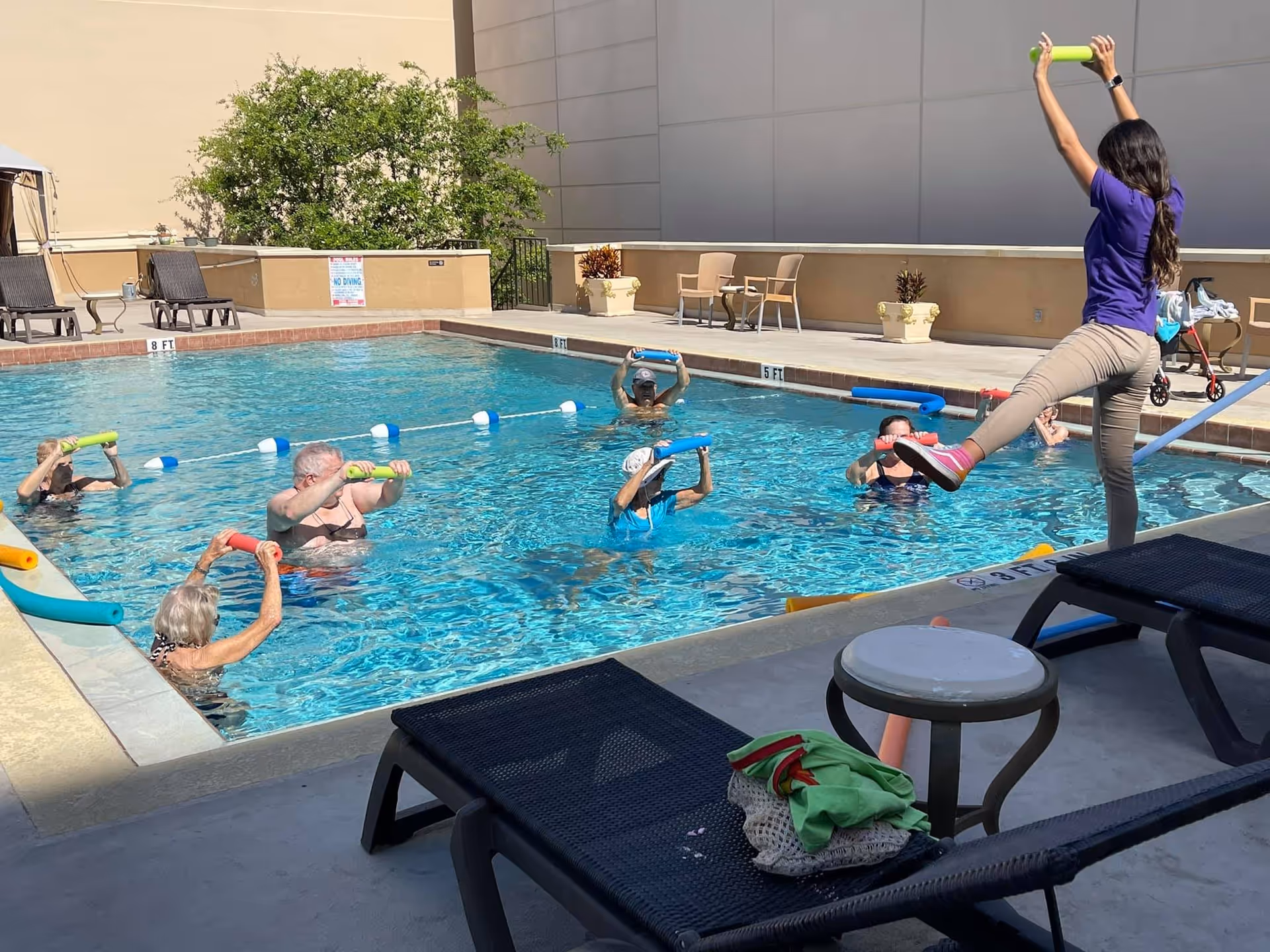 A group of elderly people participating in a water exercise class in an outdoor swimming pool, led by an instructor standing on the pool deck holding a green foam bar. The pool area has lounge chairs, potted plants, and a beige wall in the background.