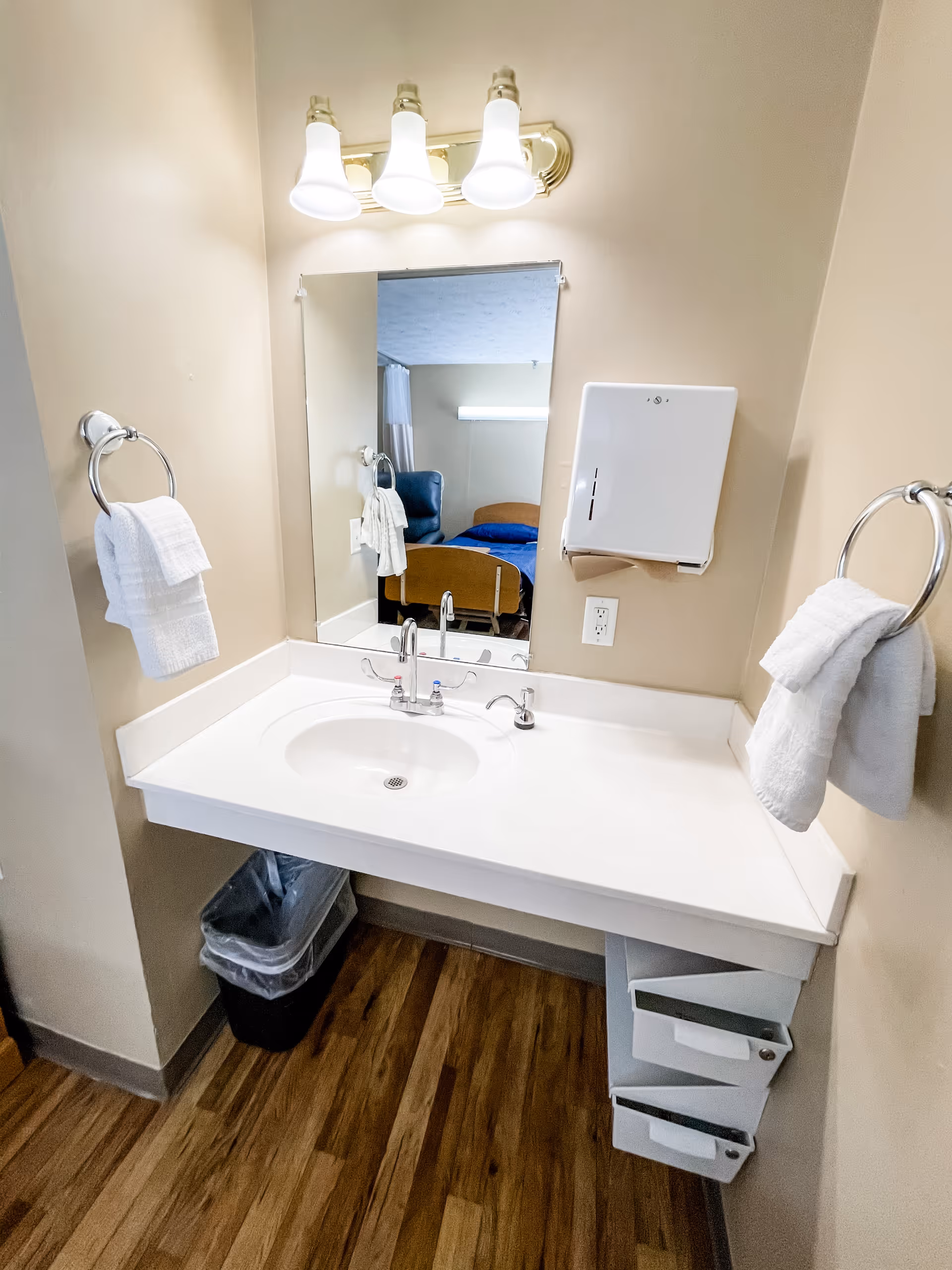 A clean bathroom sink area with a white countertop, a mirror above the sink, and three light fixtures mounted on the wall. There are two towel rings on either side of the sink, each holding white towels. A paper towel dispenser is mounted on the wall to the right of the mirror. The floor is wood-patterned, and a trash bin is placed under the sink. The reflection in the mirror shows part of a room with a bed and a chair.