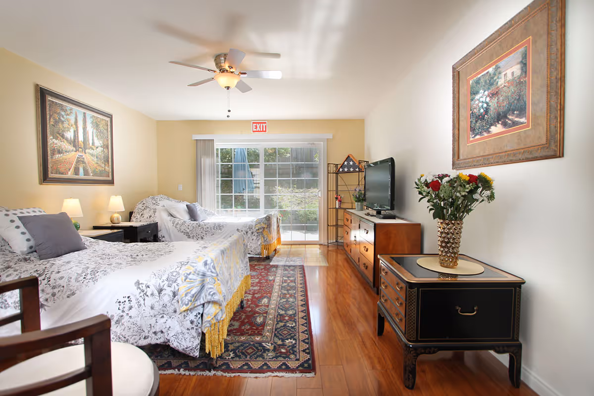 A bright and cozy bedroom with two beds covered in white and gray floral bedding. The room features wooden flooring, a ceiling fan with light, two framed paintings on the walls, a flat-screen TV on a wooden dresser, a small black side table with a vase of flowers, and a sliding glass door leading outside with an exit sign above it.
