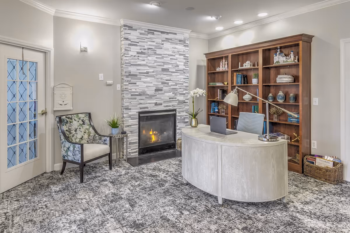 Well-lit interior sitting area with a curved reception desk, fireplace, bookshelf, and an upholstered chair.