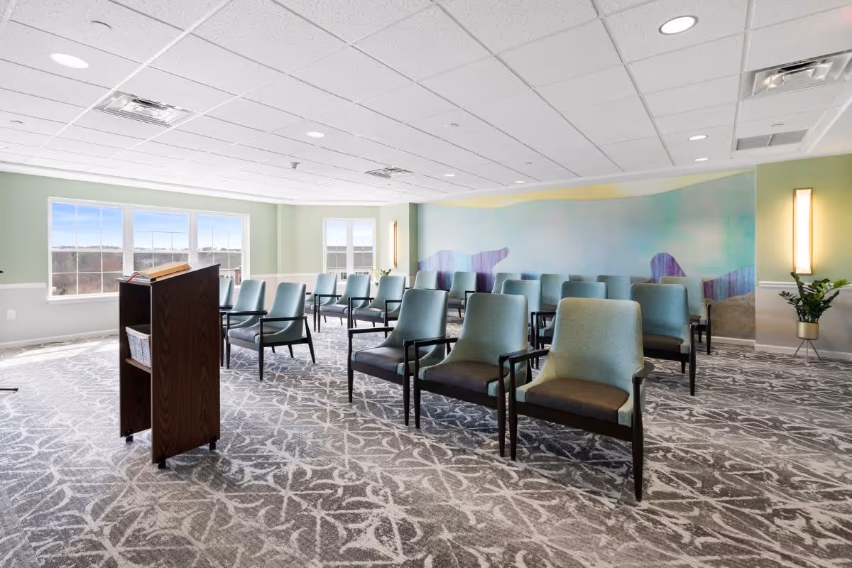 A bright, spacious room with multiple rows of blue cushioned chairs arranged facing a wooden podium. The room has large windows letting in natural light, a patterned carpet, and a colorful abstract mural on one wall. There is a potted plant near the wall with a vertical light fixture.