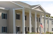 Exterior view of a two-story beige building with white columns and an American flag mounted near the entrance under a clear sky.