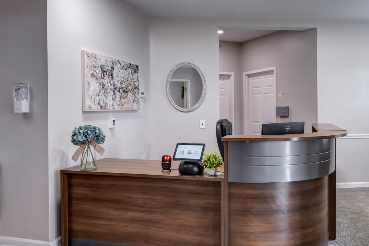 Reception desk area in a senior living facility with a wooden desk featuring a computer monitor, a tablet, a small potted plant, and a vase with blue flowers. Behind the desk is a black office chair, a round mirror on the wall, and two closed white doors. A floral painting hangs on the wall to the left, and a hand sanitizer dispenser is mounted on the wall near the corner.