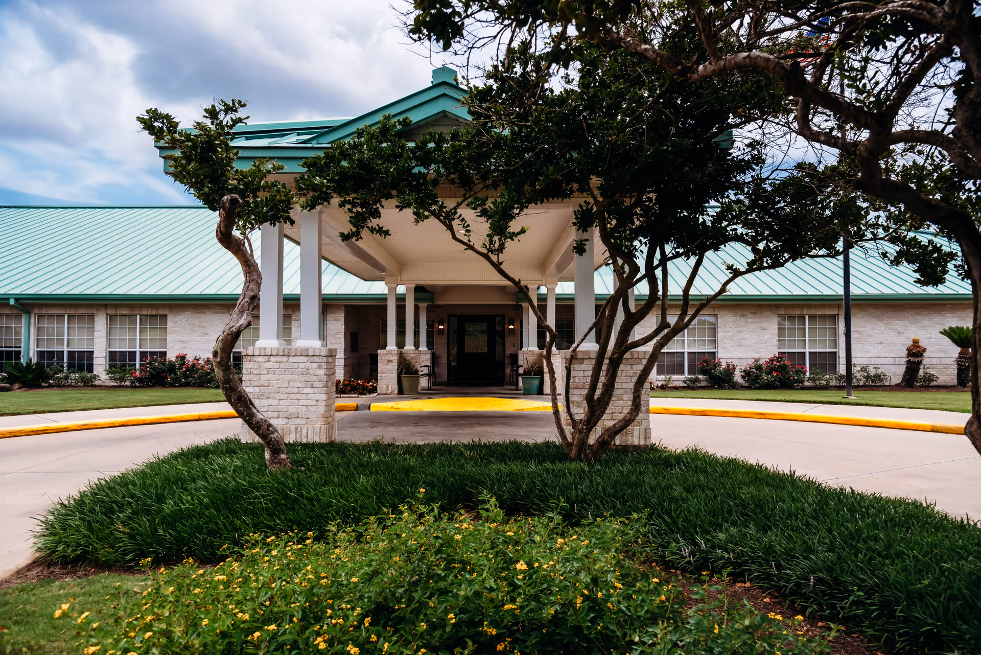 Covered main entrance of a single-story nursing facility with a porte-cochere, circular driveway, and landscaped trees and shrubs.