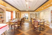 A bright dining room with multiple wooden tables and chairs arranged neatly. The room features large windows with white blinds, built-in shelves with decorative items, and a patterned ceiling. The floor has a wooden parquet design.