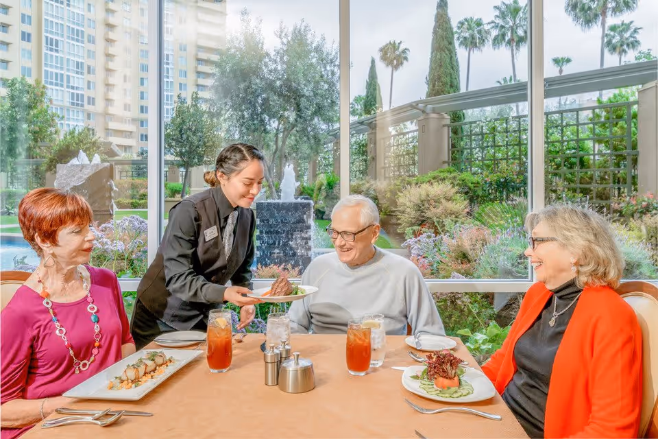 A waitress serving a plate of food to an elderly man seated at a dining table with two elderly women. The table is set with plates of food, glasses of iced tea, and silverware. Large windows behind them show a garden with trees, plants, and a water fountain.
