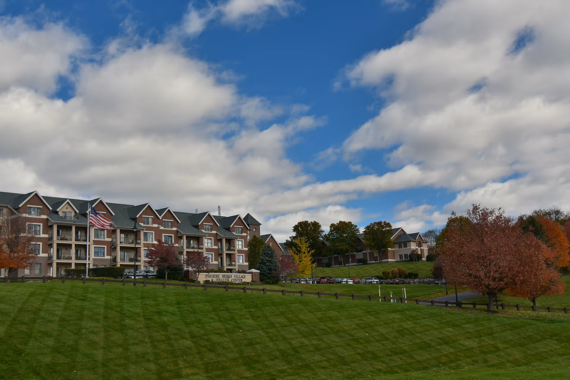 Exterior view of Phoebe Berks Health Care Center, a multi-story building with balconies, surrounded by a well-maintained green lawn and trees with autumn foliage under a partly cloudy blue sky.