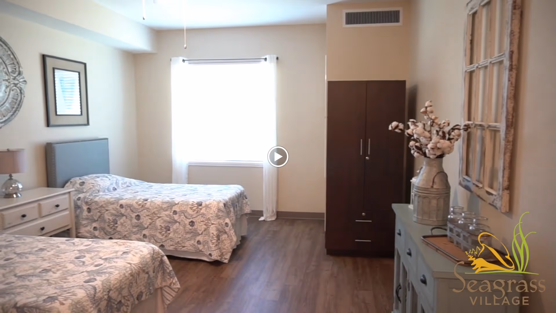 A bedroom with two twin beds covered in floral bedspreads, a nightstand with a lamp between the beds, a window with white curtains, a dark wooden wardrobe, and a dresser with decorative items including a vase with cotton stems. The room has wooden flooring and light-colored walls.