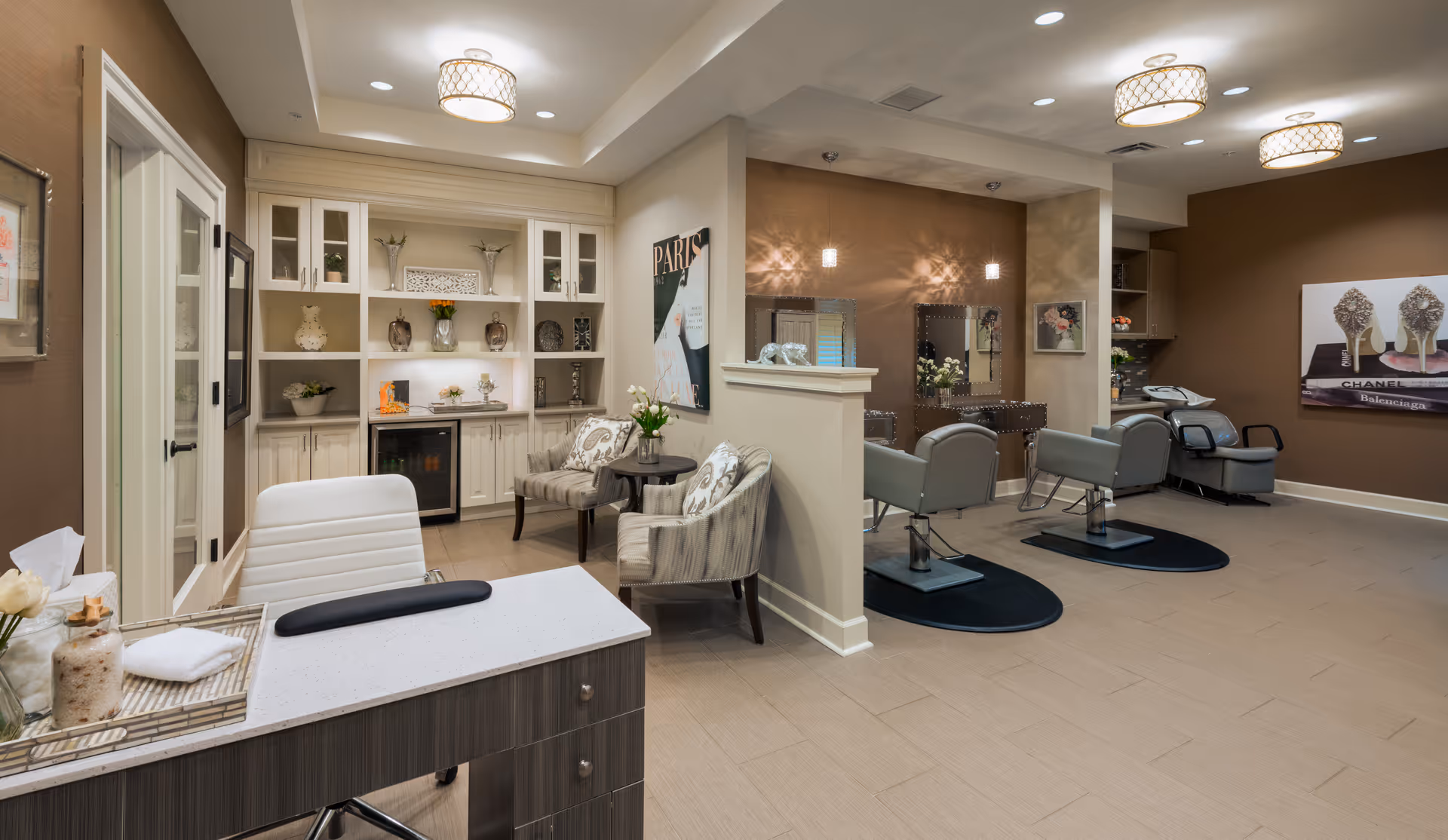 Interior view of a stylish salon area in The Kensington Reston with two salon chairs facing mirrors on a brown accent wall, a white desk with a white chair in the foreground, and a cozy seating area with two patterned armchairs and a small table. The space is decorated with shelves holding vases and decorative items, soft lighting fixtures, and artwork on the walls.