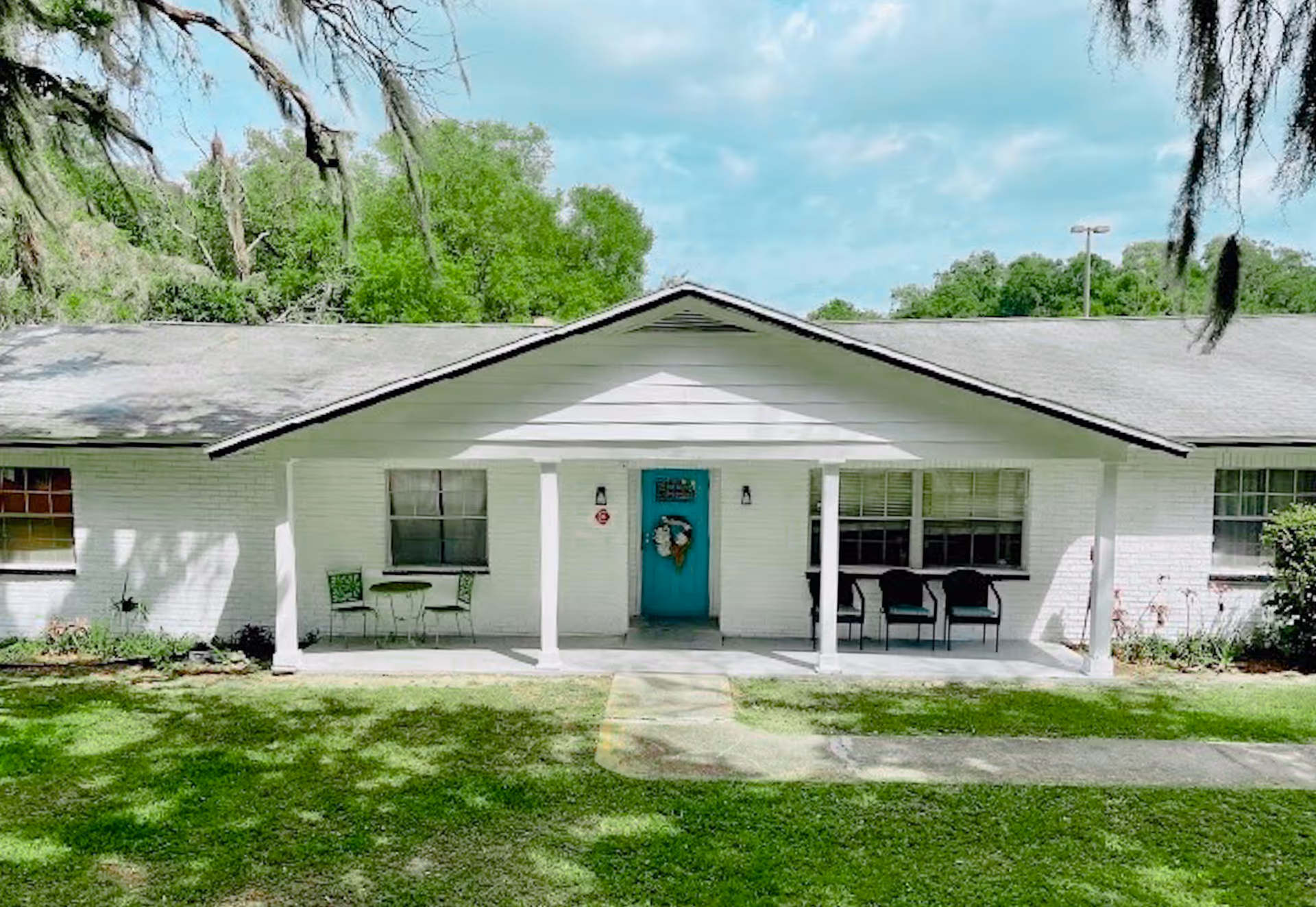 Front exterior view of a single-story white brick building with a teal door decorated with a wreath. The building has a covered porch with white pillars, two chairs and a small table on the left side, and three chairs on the right side. There is a green lawn in front and trees in the background under a partly cloudy sky.