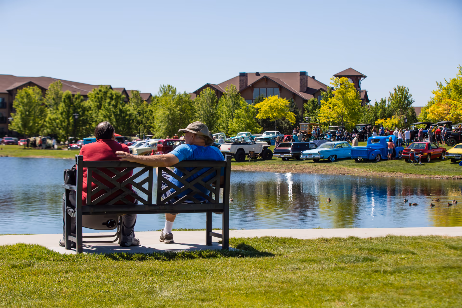 Two people sitting on a bench by a pond, with a view of a car show featuring classic cars and a large building surrounded by trees in the background.