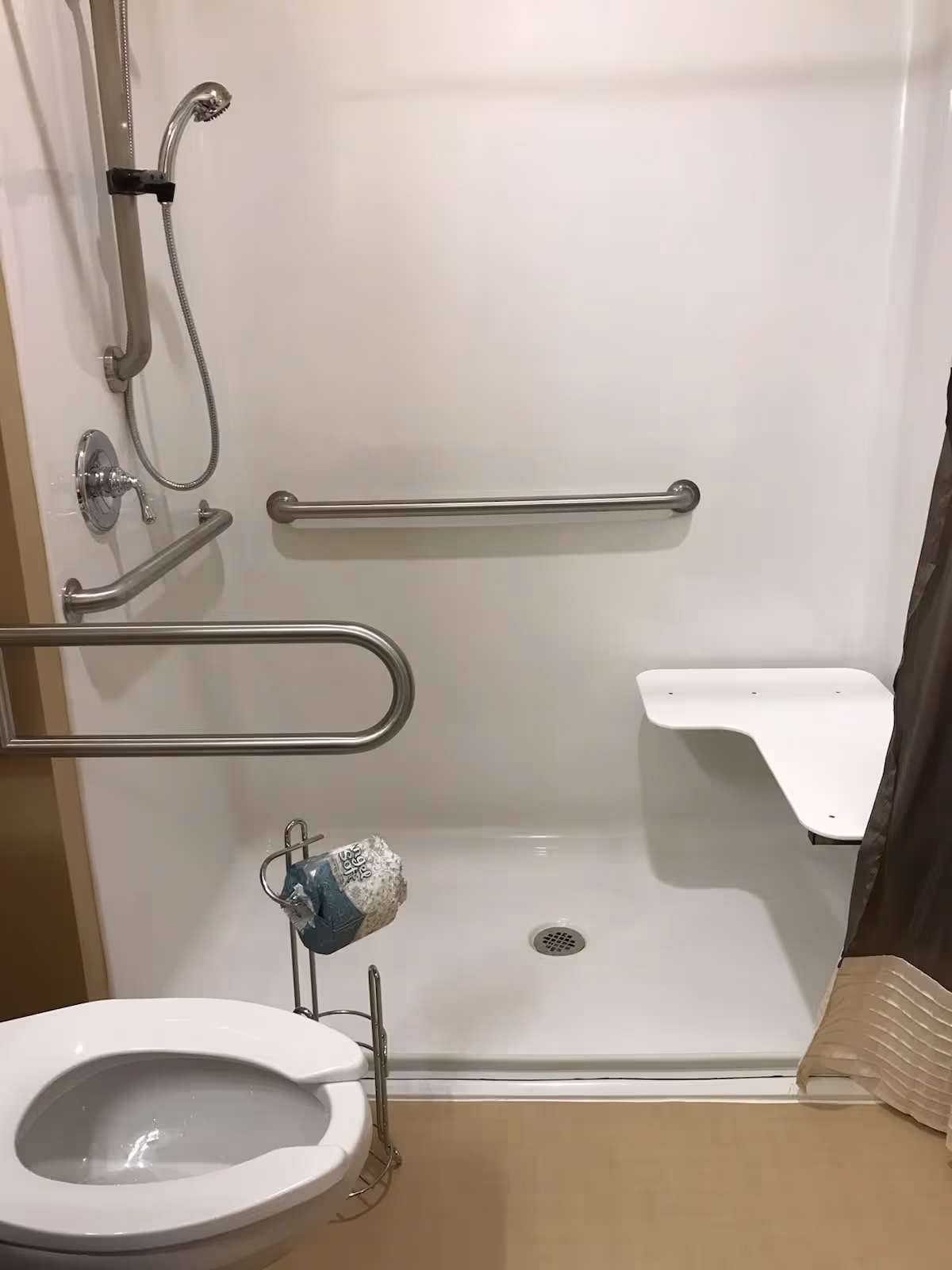 Accessible bathroom with a white toilet, a roll of toilet paper on a metal holder, a walk-in shower with a handheld showerhead, grab bars on the walls, and a fold-down shower seat. The shower curtain is partially visible on the right side.