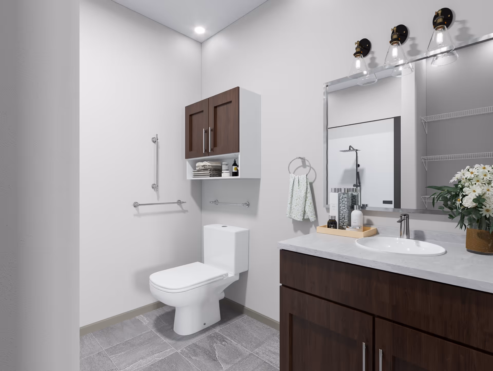 A modern bathroom with a white toilet, a dark wood vanity with a white countertop and an oval sink, a large mirror with three light fixtures above it, a wall-mounted cabinet with dark wood doors and open shelves holding towels and toiletries, a towel ring with a hanging hand towel, and a vase with white flowers on the countertop. The floor is tiled in gray, and the walls are painted white.