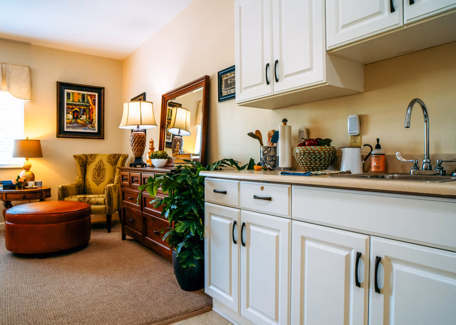 Interior view of a senior living facility room featuring a white kitchenette with cabinets, a sink, and countertop accessories including a paper towel holder and soap dispenser. Adjacent to the kitchenette is a cozy sitting area with a green patterned armchair, a round brown ottoman, a wooden dresser with a table lamp and a large mirror, a potted plant, and framed artwork on the wall.