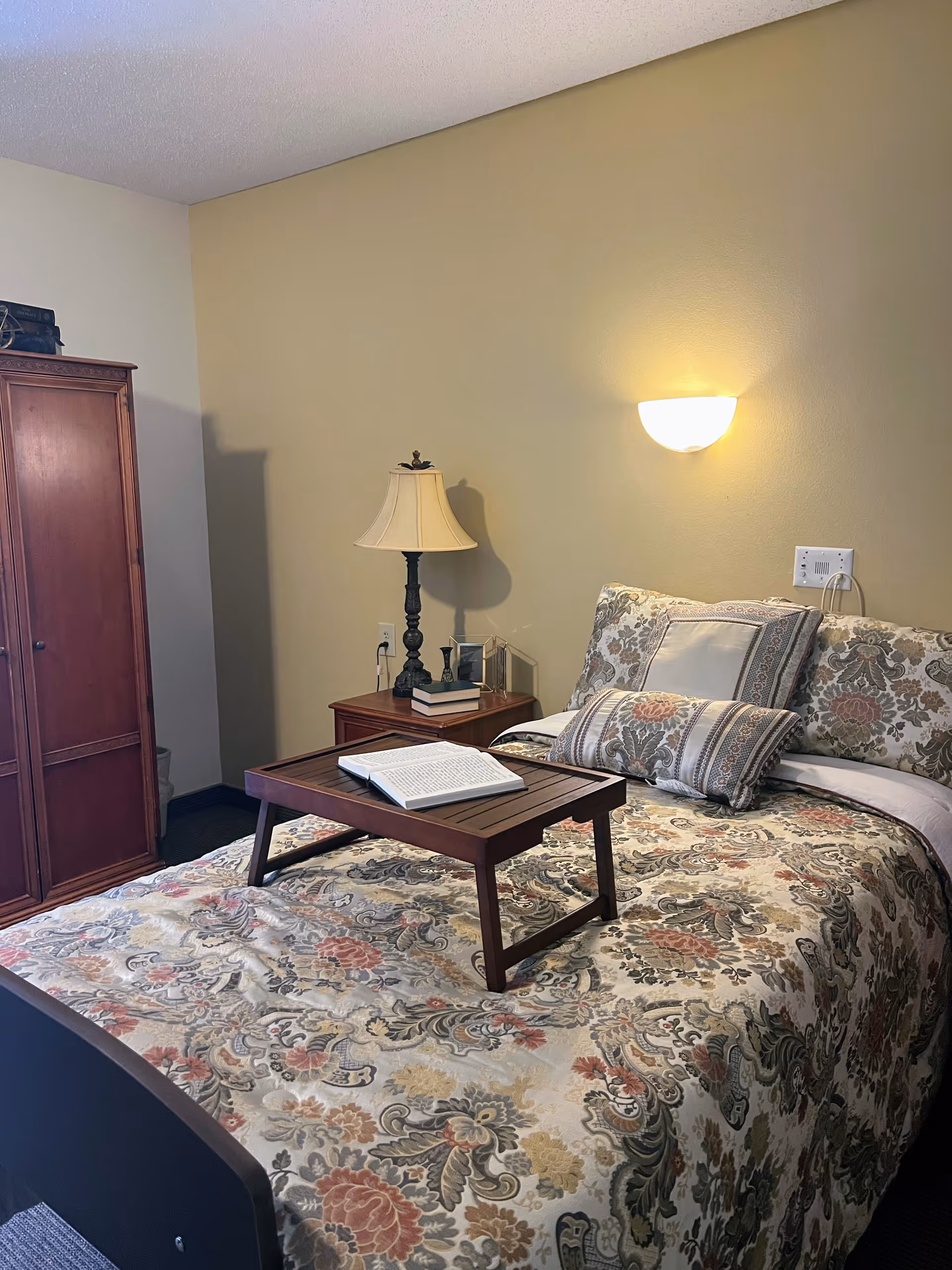 A cozy bedroom with a floral patterned bedspread and matching pillows. A wooden tray with an open book rests on the bed. Next to the bed is a wooden nightstand with a classic table lamp, a couple of books, and a small decorative item. A wooden wardrobe stands against the wall, and a wall-mounted light fixture illuminates the room.