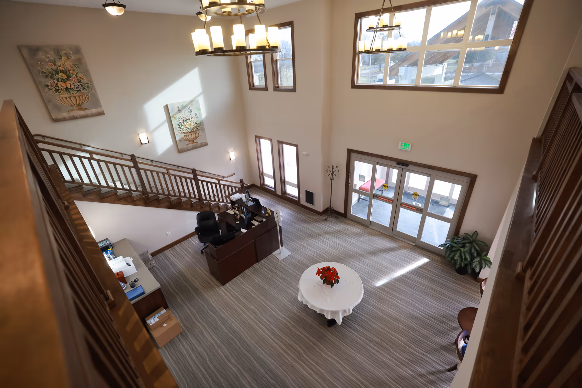 View down into a two-story senior living lobby with a reception desk, staircase, round table with flowers, and glass double doors.