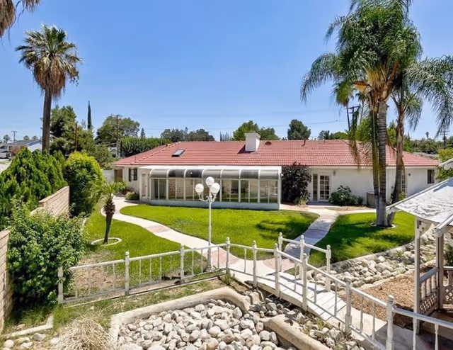 Single-story red-roofed building overlooking a landscaped backyard with palm trees, a lawn, walkways, a decorative bridge and a gazebo.
